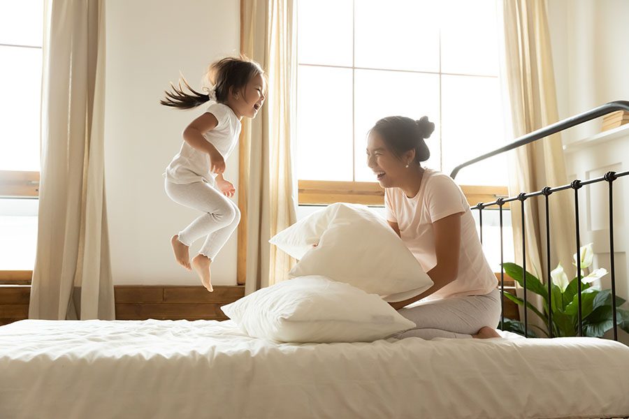 Blog - Mother Sitting on the Bed Laughing as she Watches Her Cheerful Daughter Jumping on the Bed